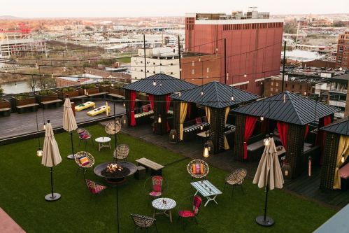 Rooftop lounge with cabanas, seating areas, and a fire pit, overlooking a cityscape at sunset.