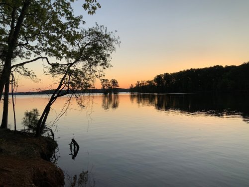 A serene lake at sunset, with calm waters reflecting the colorful sky and silhouettes of trees along the shore.