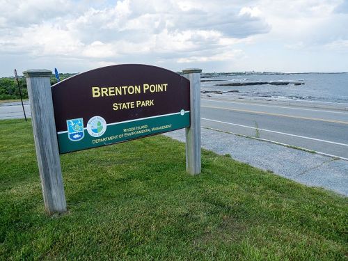 Sign for Brenton Point State Park near the ocean, with grassy area and cloudy sky in the background.