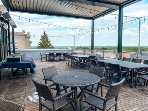 Rooftop patio with tables and chairs, string lights, and a view of greenery under a partly cloudy sky.
