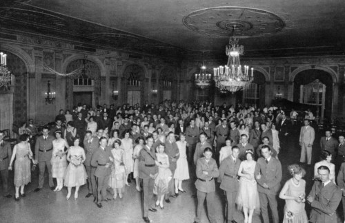 A large group of elegantly dressed people in a grand hall, with chandeliers and a piano in the background.