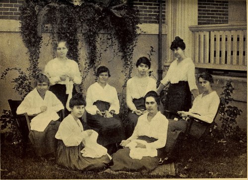 A group of eight women in historical clothing, seated and standing outdoors, surrounded by greenery and a building.