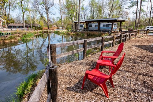 Two red chairs by a pond, with a wooden fence and campers in the background surrounded by trees.