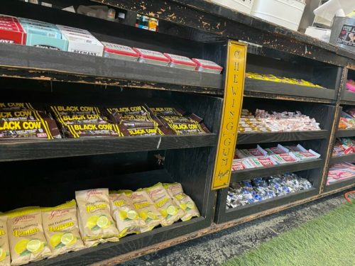 A display of various candies and chewing gum in a wooden shelf, featuring "Black Cow" and "Lemon" flavors.