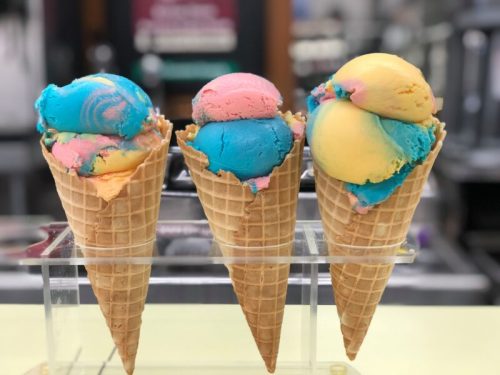 Three ice cream cones with colorful scoops: pink, blue, yellow, and orange, displayed on a clear stand.