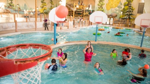 A lively indoor water park with children playing in the pool, basketball hoops, and colorful inflatable floats.