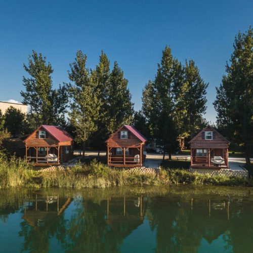 Three wooden cabins by a calm lake, surrounded by tall trees and reflecting in the water.