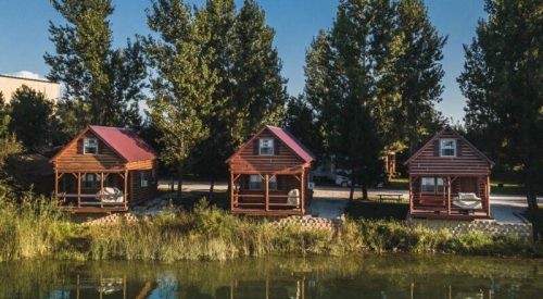 Three wooden cabins with porches beside a calm pond, surrounded by tall trees under a clear blue sky.