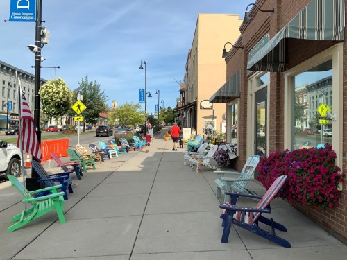 Colorful Adirondack chairs line a sidewalk in a quaint town, with shops and flowers in the background on a sunny day.