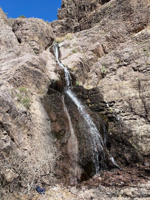 A small waterfall cascading down rocky terrain, surrounded by dry vegetation and steep cliffs under a clear blue sky.