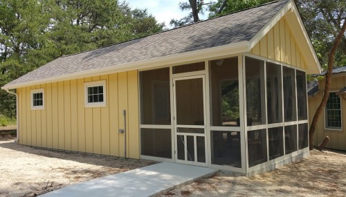 A yellow cabin with a screened porch, surrounded by trees and a gravel path leading to the entrance.