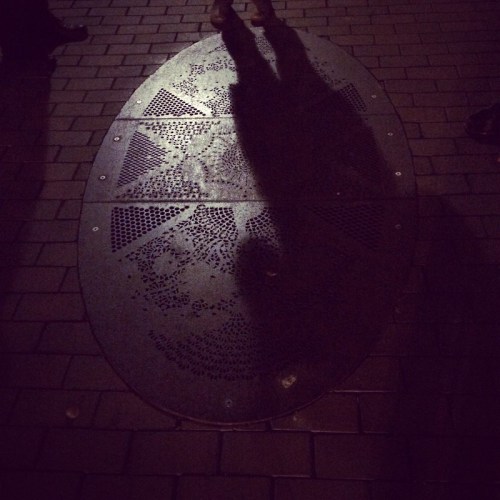 A dark street scene featuring a patterned manhole cover with shadows of people standing nearby.