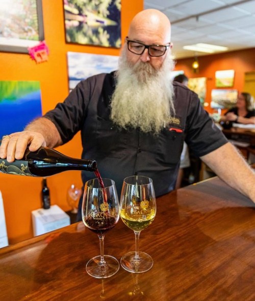 A bearded man pours red wine into a glass, with a second glass of white wine on a wooden bar. Colorful artwork is in the background.