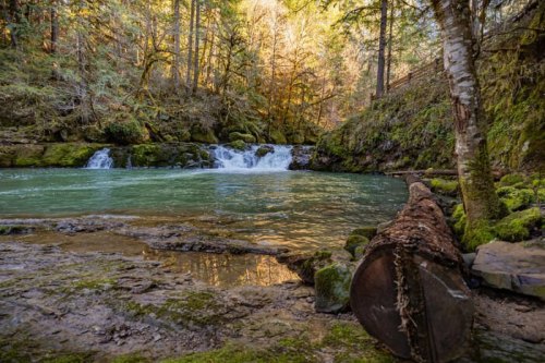 A serene river scene with gentle waterfalls, surrounded by lush greenery and autumn-colored trees.