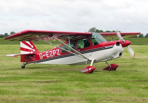 A red and white light aircraft with the registration G-EZPZ parked on a grassy airfield under a cloudy sky.