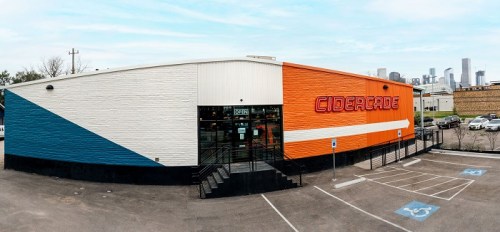 Exterior view of Cidercade, featuring colorful walls and a city skyline in the background. Accessible parking visible.