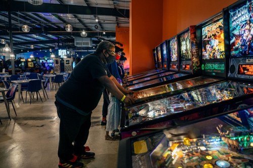 Three people play pinball machines in a vibrant arcade filled with colorful lights and games.