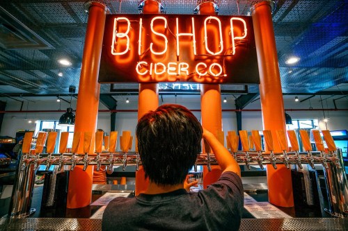 A person stands in front of a bar with multiple cider taps, under a glowing "Bishop Cider Co." neon sign.