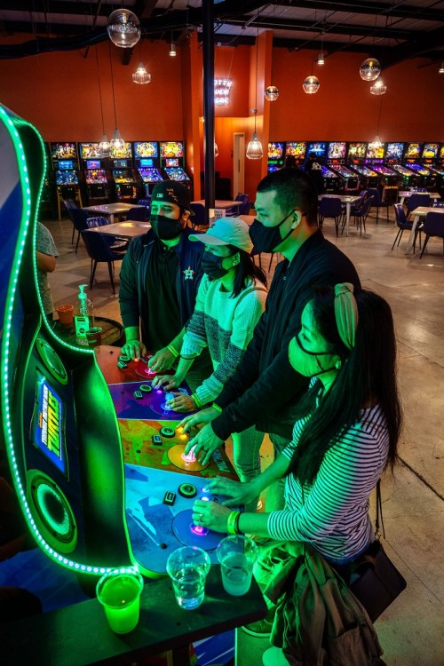 Four people wearing masks play an arcade game in a vibrant gaming venue with colorful lights and machines in the background.