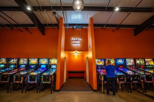A vibrant arcade with colorful pinball machines and a sign reading "Potty Over Here" on the wall.