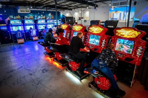 Four people playing racing arcade games in a vibrant gaming arcade with colorful machines in the background.