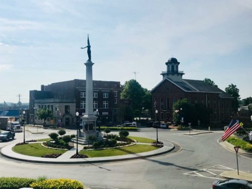 A town square featuring a monument, a church, and surrounding buildings, with flowers and an American flag in view.