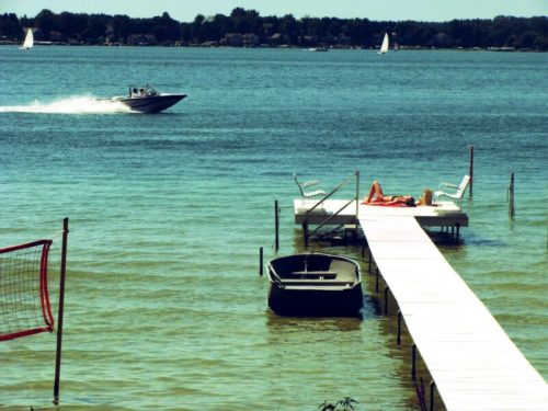 A serene lake scene with a dock, a boat speeding by, and a person relaxing on the dock under the sun.