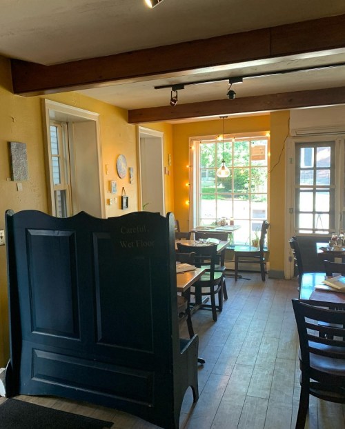 Cozy restaurant interior with wooden floors, yellow walls, and tables by a large window. Sign reads "Careful, Wet Floor."