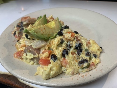 A plate of scrambled eggs with black beans, tomatoes, and avocado slices, garnished with spices.