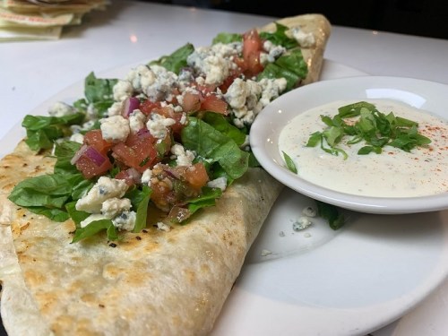 A large flatbread filled with lettuce, tomatoes, and blue cheese, served with a side of creamy dressing.