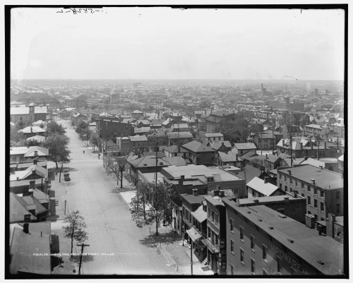 Aerial view of a cityscape featuring rows of houses, streets, and distant buildings in black and white.
