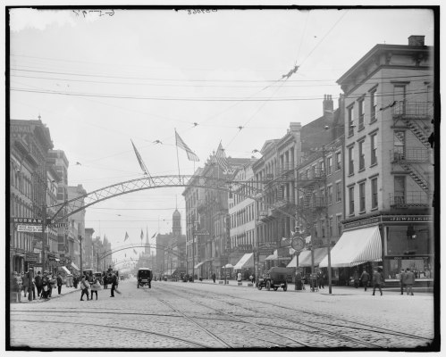 Historic street scene with buildings, flags, and horse-drawn carriages under an archway, capturing early 20th-century urban life.