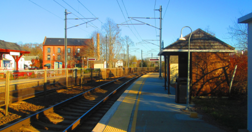 A quiet train station platform with tracks, a small shelter, and buildings in the background under a clear blue sky.
