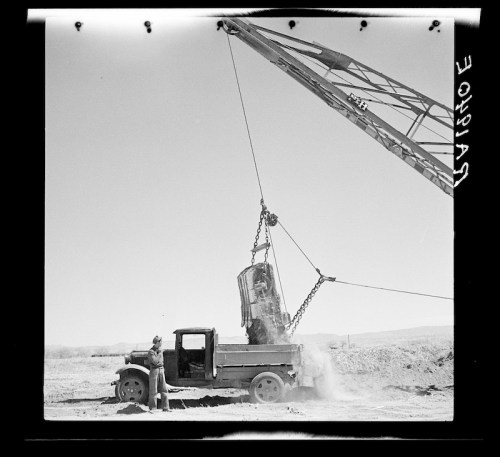 A vintage truck with a crane lifting a heavy object in a dusty landscape under a clear sky.