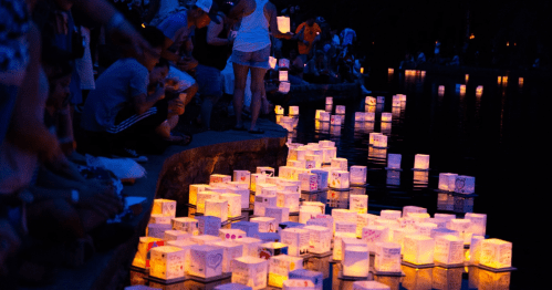 A serene night scene with glowing lanterns floating on water, surrounded by people enjoying the ambiance.