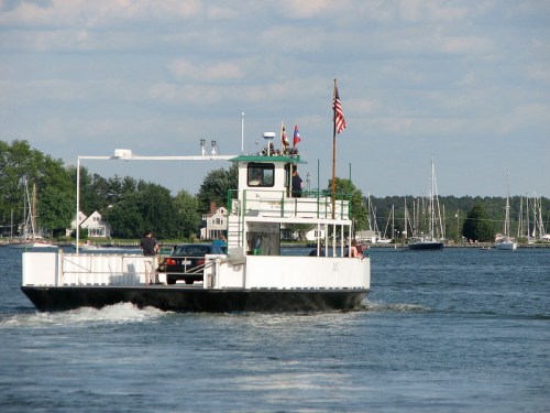A ferry boat carrying a car navigates a calm waterway, with sailboats and trees in the background under a blue sky.
