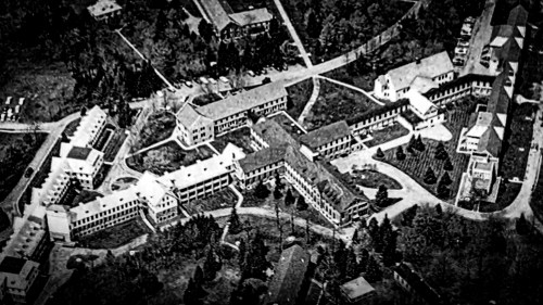 Aerial view of a large, complex building layout surrounded by trees and pathways, in black and white.