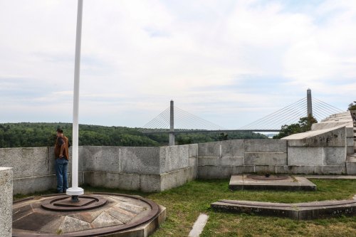 A person stands on a stone platform overlooking a bridge and lush green landscape under a cloudy sky.