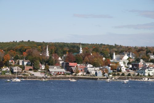 A scenic view of a coastal town with colorful autumn trees, churches, and boats on the water under a clear blue sky.