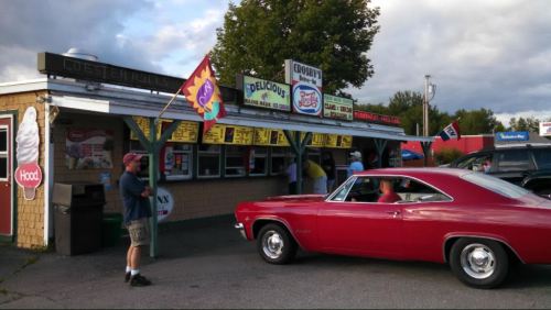 A vintage red car parked outside a retro ice cream stand with colorful signs and a flag, surrounded by trees.