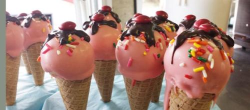 Colorful ice cream cones with pink scoops, chocolate drizzle, and sprinkles, topped with red candies.