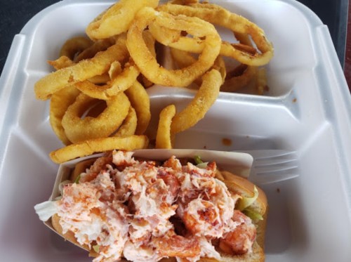 A takeout container with a lobster roll topped with shredded lobster meat and a side of crispy onion rings.
