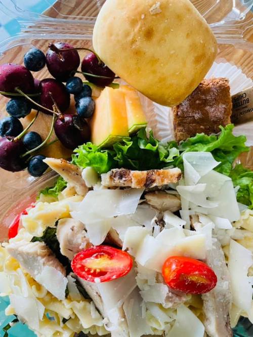 A colorful salad with chicken, pasta, cherry tomatoes, greens, fruit, a roll, and a dessert in a clear container.