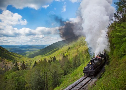 A steam train chugs along a scenic mountain track, billowing smoke against a backdrop of lush green hills and blue skies.
