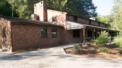 A brick house with a sloped roof, chimney, and porch, surrounded by trees and a gravel driveway.