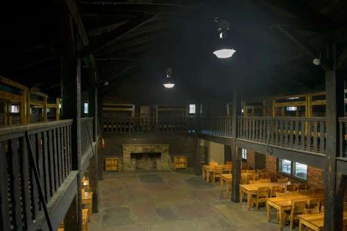 Interior of a rustic hall with wooden beams, tables, and a stone fireplace, featuring a two-level seating arrangement.