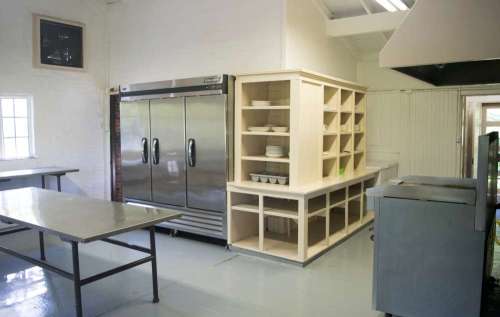 A spacious kitchen with stainless steel refrigerators, wooden shelving, and tables for food preparation.