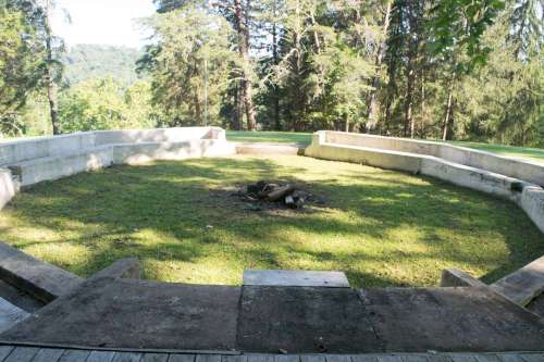 A circular stone fire pit area surrounded by grass and trees, with a view of a wooded landscape in the background.