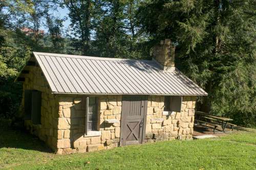 A small stone cabin with a metal roof, surrounded by trees and a picnic table nearby.