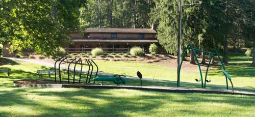 A playground with swings and climbing equipment, set in a grassy area near a building surrounded by trees.